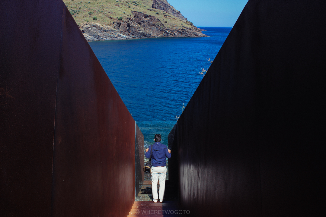 Person standing in a narrow gap between dark walls framing a view of the sea and a headland